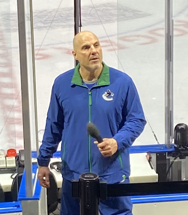 Ein Mann in einer blauen Jacke hält ein Mikrofon, während er vor einer Eishockeyarena steht, mit einer Glaswand und Equipment im Hintergrund.