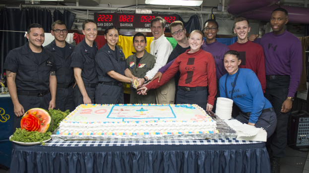 Eine Gruppe strahlender Menschen um einen Tisch mit einem Kuchen, Lebensmitteln und einer Wassermelone versammelt, die ein besonderes Ereignis in einem Raum mit Vorhängen, einer digitalen Anzeigetafel, Lampen, Rohren und einer Wand feiern.