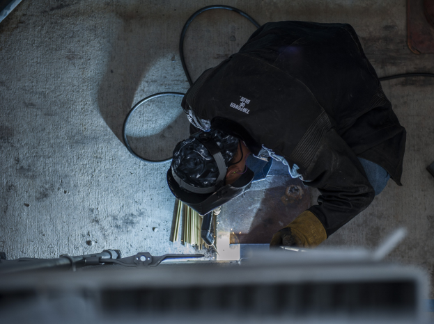 Ein Schweißer in Schutzausrüstung verwendet eine Schweißmaschine an einem Metallstück, umgeben von Werkzeugen und Gegenständen auf dem Boden.