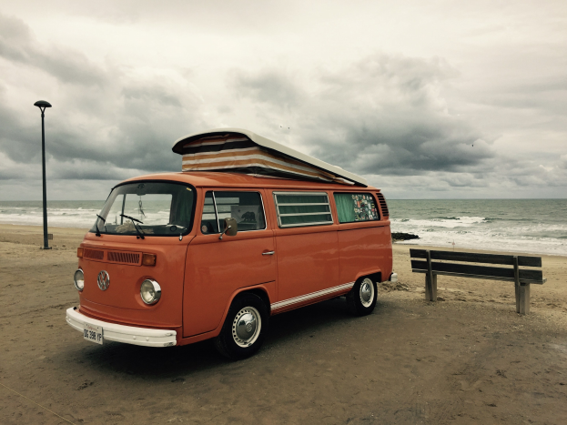 Oranger VW-Bus am Strand bei der See geparkt, mit einer Bank und Laterne in der N├Ąhe, unter einem bew├Âlktem Himmel mit sichtbarem Wasser im Hintergrund.
