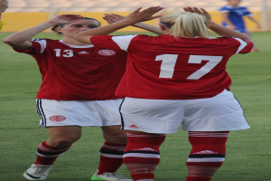 Zwei Frauen in roten T-Shirts und weißen Shorts stehen auf einem Fußballfeld und klatschen begeistert in die Hände, mit einer Person in einem blauen Kleid, einer gelben Tafel und ein paar Stühlen im Hintergrund.