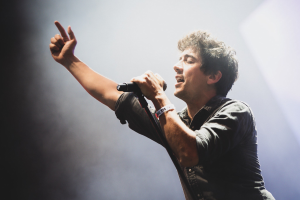 Ein Mann in schwarzem Hemd und Jeans singt leidenschaftlich in ein Mikrofon auf einer hell erleuchteten Bühne, seine Augen geschlossen, während das Publikum im Hintergrund applaudiert.