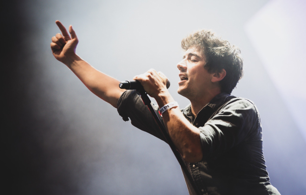 Ein Mann in schwarzem Hemd und Jeans singt leidenschaftlich in ein Mikrofon auf einer hell erleuchteten Bühne, seine Augen geschlossen, während das Publikum im Hintergrund applaudiert.