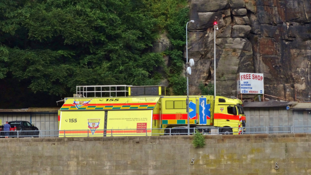Ambulanz am Rande einer Straße neben einer Klippe geparkt, mit verschiedenen Gegenständen wie einer Wand, einem Geländer, Masten, Lichtern und Schildern mit Text im Vordergrund und Bäumen und einem Auto im Hintergrund.