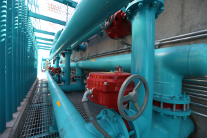 Eine gro├če Gruppe von blauen Rohren und Ventilen in einem Geb├Ąude, mit einem prominenten roten Ventil in der Mitte, Metallst├Ąben auf der linken Seite und einer Wand im Hintergrund, die wahrscheinlich den Prozess des Bauens einer Gasleitung darstellt.