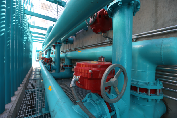 Eine gro├če Gruppe von blauen Rohren und Ventilen in einem Geb├Ąude, mit einem prominenten roten Ventil in der Mitte, Metallst├Ąben auf der linken Seite und einer Wand im Hintergrund, die wahrscheinlich den Prozess des Bauens einer Gasleitung darstellt.