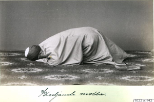 Ein altes Schwarz-Weiß-Foto eines Mannes im Gebet, der ein Tuch und eine Kappe trägt, mit der Inschrift "Bredjande Molla, die erste muslimische Frau der Welt" unten.
