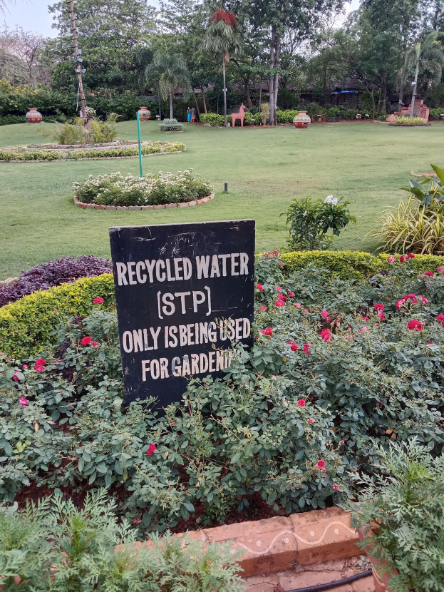 Schilder lesend "Recyceltes Wasser (STP) nur für Garten" in einem Garten mit grünem Gras, bunten Blumen, hohen Bäumen und einem klaren blauen Himmel.