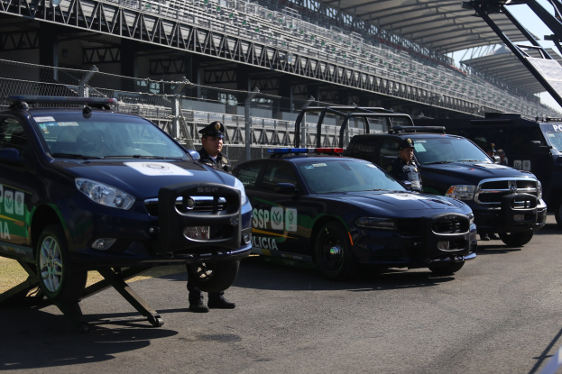 Eine Gruppe von Polizeiwagen vor einer Rennstrecke geparkt, uniformierte Beamte stehen auf der Straße, im Hintergrund ein Stadion und Maschendrahtzaun.