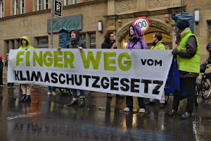 Eine Gruppe von Menschen steht auf einer Straße und hält ein Schild hoch, auf dem "Finger Weg Klimaschutzgesetz" steht, im Protest. Im Hintergrund sind ein Gebäude, ein Schild und ein Fahrrad zu sehen.