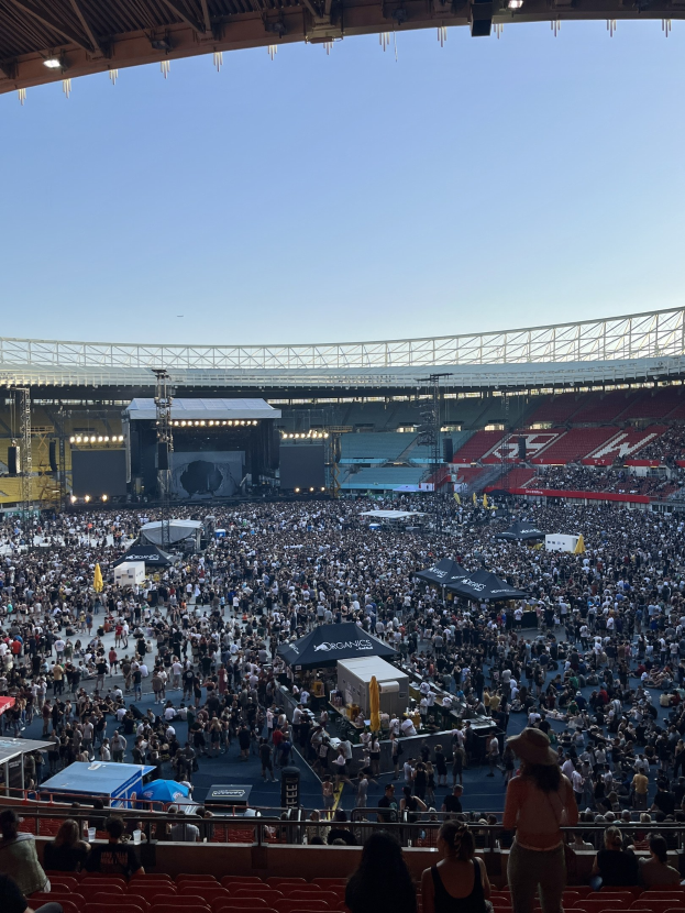 Eine große Menschenmenge sitzt und steht in einem Stadion mit Geländern, Zelten und Lichtern unter einer Decke mit Beleuchtung, mit dem Himmel im Hintergrund.