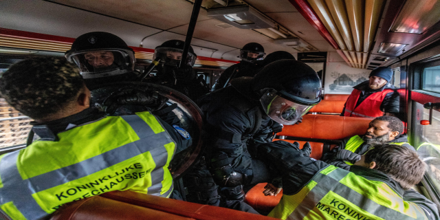 Eine Gruppe von Polizisten in Einsatzausrüstung auf einem Bus, einige tragen Helme und Westen, mit einer Person in der Mitte und einem Plakat im Hintergrund.