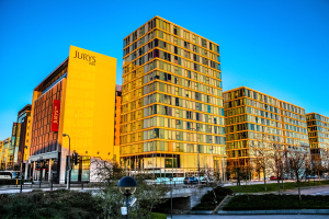 Eine belebte Stadtlandschaft mit hohen Gebäuden, darunter das Jurys Inn Birmingham City Centre, mit Kraftfahrzeugen auf der Straße, Straßenmasten, Laternen und Signalen, umgeben von Büschen und Bäumen unter einem klaren blauen Himmel.