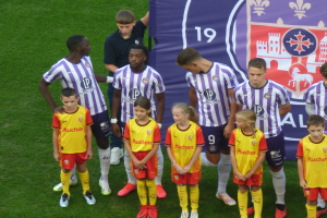 Eine Gruppe junger Menschen auf einem saftig grünen Fussballfeld, mit einem Banner im Hintergrund, das 'Ligue 1' lautet, einige tragen Trikots und andere Shorts, unter einem hellblauen Himmel.