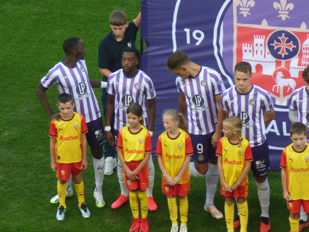 Eine Gruppe junger Menschen auf einem saftig grünen Fussballfeld, mit einem Banner im Hintergrund, das 'Ligue 1' lautet, einige tragen Trikots und andere Shorts, unter einem hellblauen Himmel.
