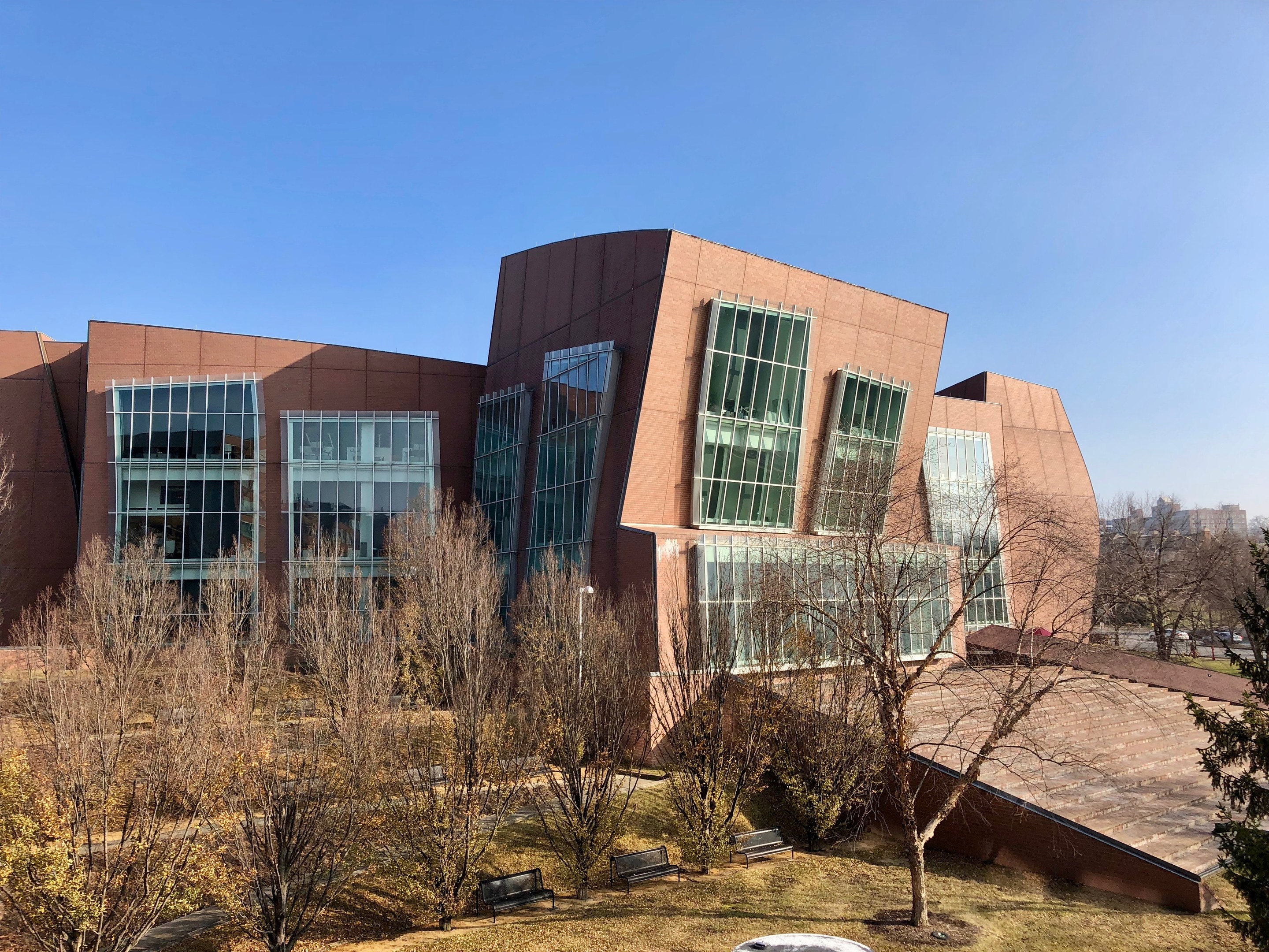 Großes mehrstöckiges Gebäude mit zahlreichen Fenstern, umgeben von Bäumen, Bänken, Gras und einem Lichtmast, mit anderen Gebäuden und einem klaren blauen Himmel im Hintergrund.