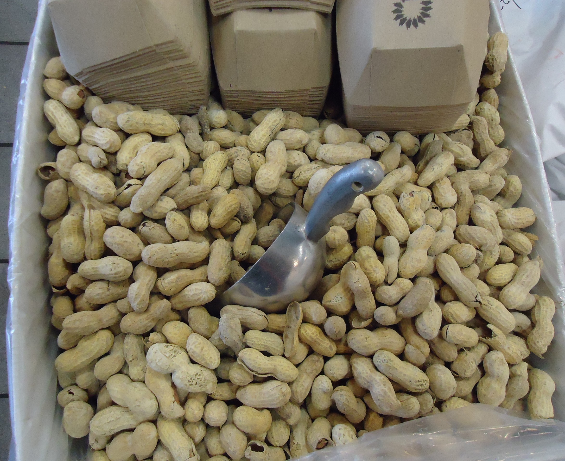 Eine Schachtel Erdnüsse auf einem Tisch mit einer Plastikabdeckung und einem Löffel daneben, umgeben von zusätzlichen Schachteln Erdnüssen.