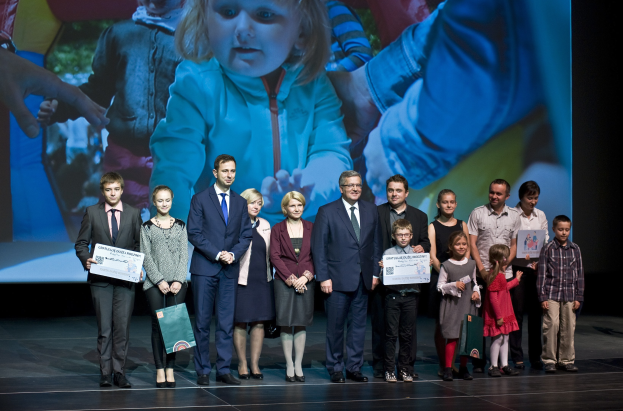 Eine Gruppe von Menschen auf einer Bühne, einige halten Taschen und Urkunden, im Hintergrund ein Bildschirm mit einem Bild von einem Kind und anderen Menschen.