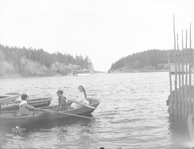 Ein Schwarz-Weiß-Foto von drei Menschen in einem Boot auf einem See, wobei einer paddelt und die anderen beiden sitzen, mit Pfählen auf der rechten Seite und einem weiteren Boot und Bäumen im Hintergrund.