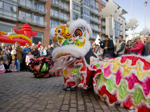 Ein lebendiges chinesisches Neujahrsfest in Amsterdam mit einer Löwen-Tanzvorstellung vor einer Zuschauermenge, darunter einige, die Fotos machen, vor einer Kulisse aus Gebäuden, Laternenmasten und einem klaren blauen Himmel.