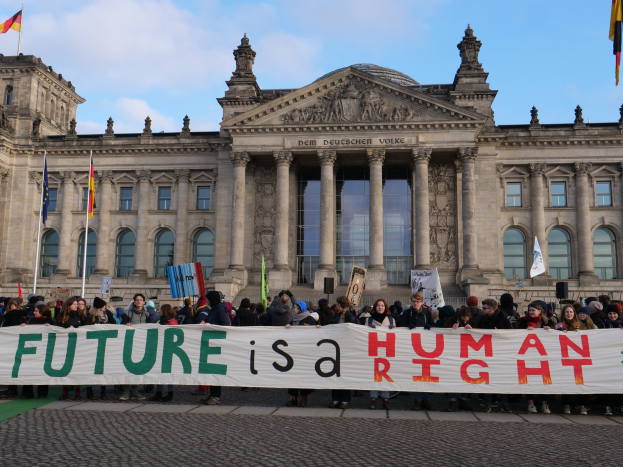 Eine Gruppe von Menschen hält ein Transparent mit der Aufschrift 'Zukunft ist ein Menschenrecht' vor dem Reichstagsgebäude in Berlin, Deutschland, mit Himmel und Fahnen im Hintergrund.