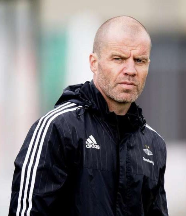 Kahlköpfiger Trainer in marineblauer Jacke mit weißen Streifen und weißem Kragen auf einem Fuballfeld mit verschränkten Händen und ernstem Gesichtsausdruck.