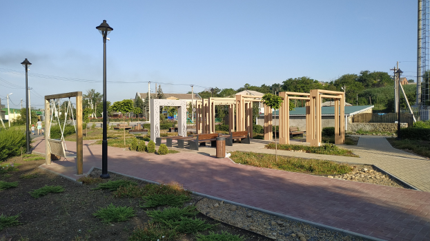 Parkszene mit einem Weg, der von hölzernen Pfählen, Bänken, Pflanzen, Gras, Steinen, Straßenlaternen, Strommasten mit Drähten, Bäumen, Gebäuden und einem Turm im Hintergrund gesäumt ist, unter einem klaren blauen Himmel.