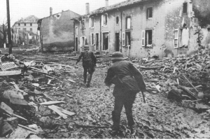 Zwei Soldaten in Helmen und mit Taschen gehen durch die Trümmer eines zerstörten Gebäudes, mit anderen Gebäuden und Pfählen im Hintergrund unter einem klaren Himmel, dargestellt in Schwarz-Weiß.