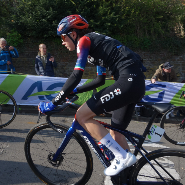Professionelle Radrennfahrerin Katie Archibald trägt einen Helm, eine Brille, Handschuhe und Schuhe und fährt ein Fahrrad auf einer Straße vorbei an einer Zuschauermenge, mit einem Banner, einer Wand, Bäumen und einem Gebäude im Hintergrund.