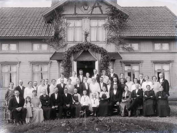Schwarzes und weißes Foto einer großen Gruppe von Menschen, die vor einem Haus mit reichlicher Vegetation und sichtbarem Himmel posieren.
