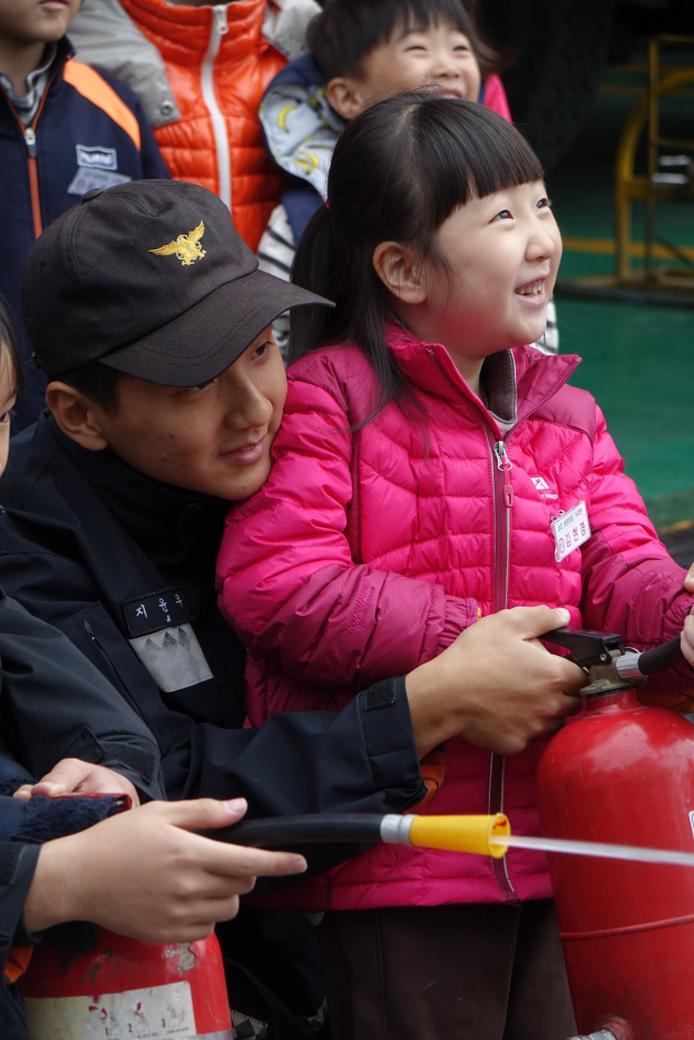 Kinder in einer Schulumgebung versammeln sich um einen roten Hydranten, einer hält ein Rohr, mit verschiedenen Gegenständen im Hintergrund.