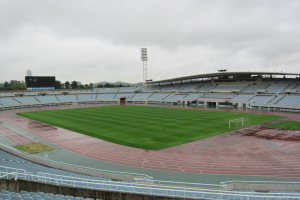 Großes Stadion mit einem Fußballfeld, umgeben von Sitzplätzen, einem Zaun, einem Anzeigebildschirm, Bäumen, einem Turm, Gebäuden und einem bewölkten Himmel.