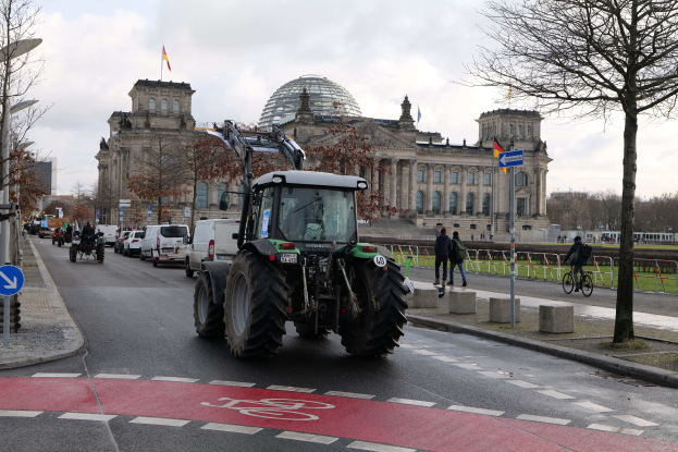 Ein Traktor fährt auf der Straße vor dem Reichstagsgebäude in Berlin, Deutschland, vorbei, während Menschen zu Fuß gehen und Fahrräder fahren, Bäume, Laternenpfähle und Schilder an der Straße stehen und das Gebäude mit Flaggen geschmückt ist.