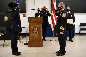 Eine Gruppe uniformierter Personen in Masken steht vor einem Podium mit Mikrofon, umgeben von Fahnen, Stühlen und Tischen, während einer Vereidigung eines neuen Feuerwehrchefs, wobei eine Person ein Zertifikat hält.