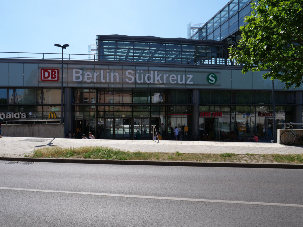 Großes gläsernes Berliner Südkreuz-Bahnhof in Deutschland mit Text an der Fassade, umgeben von Straßenlaternen, Lampen, Fahrzeugen, Fußgängern, Fahrrädern, Bäumen und einem klaren blauen Himmel.