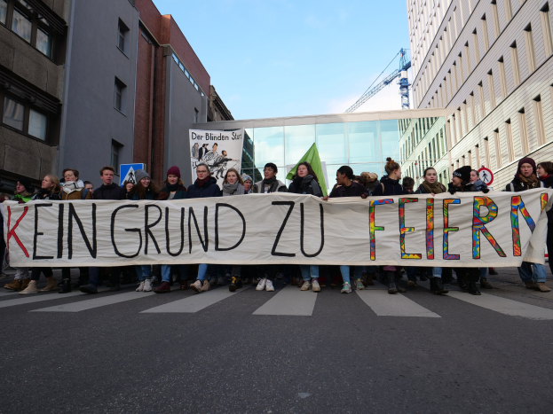 Demonstranten mit einem Transparent 'Kein Grund zu Feiern' gegen deutsche Sparmaßnahmen vor Gebäuden, einem Schild, einem weiteren Transparent, einem Kran und einem bewölkten Himmel.
