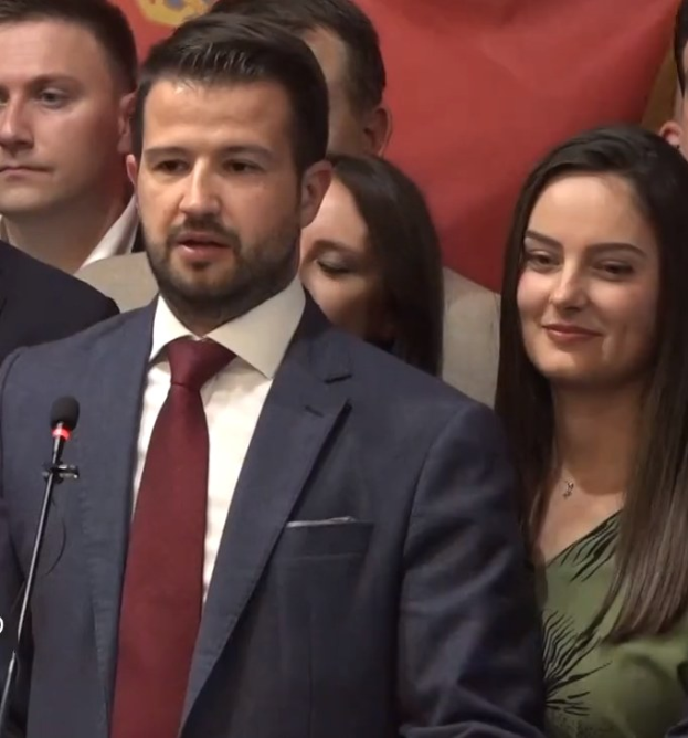 Ein Mann im Anzug spricht in ein Mikrofon an einem Podium, mit einem Banner dahinter und einer Menge davor, wahrscheinlich zu einem Regierungsverbot von sozialen Medien.