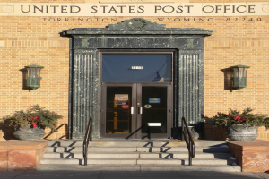 Die Poststelle der Vereinigten Staaten in Torrington, Wyoming, mit einer Tür, Treppen mit Geländern, Topfpflanzen, an der Wand montierten Leuchten und Text auf dem Gebäude.