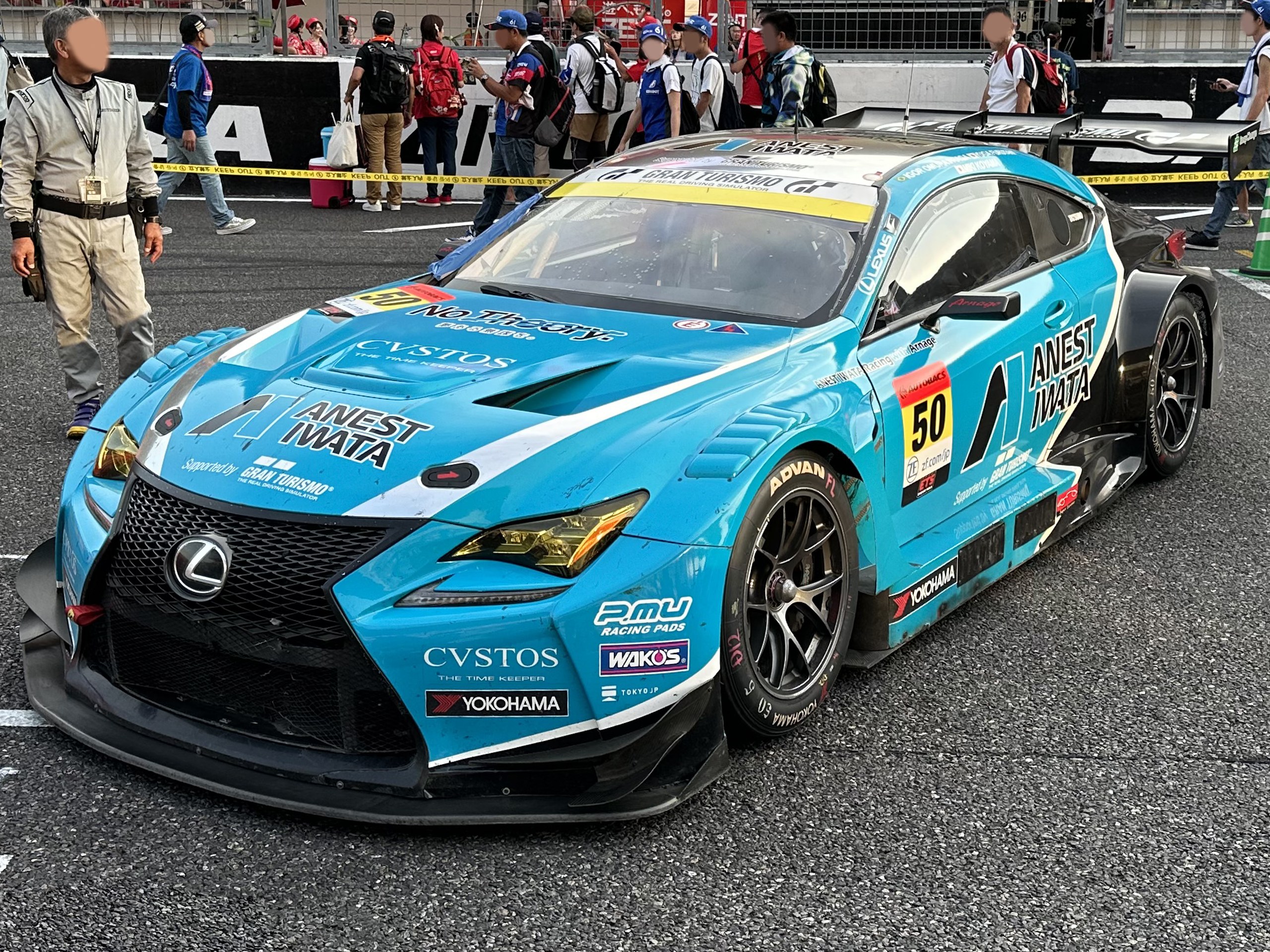 Blauer Lexus RC F GT3 Rennwagen auf einer Straße mit Zuschauern, einem Verkehrskegel rechts und Hintergrundwerbung und Gelöndern.