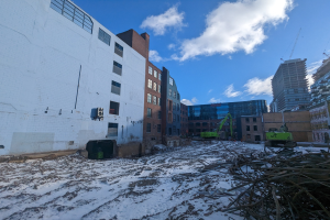 Großes Gebäude mit schneebedeckter Erde, umgeben von Fahrzeugen und Schutt, mit bewölktem Himmel und anderen Gebäuden im Hintergrund.