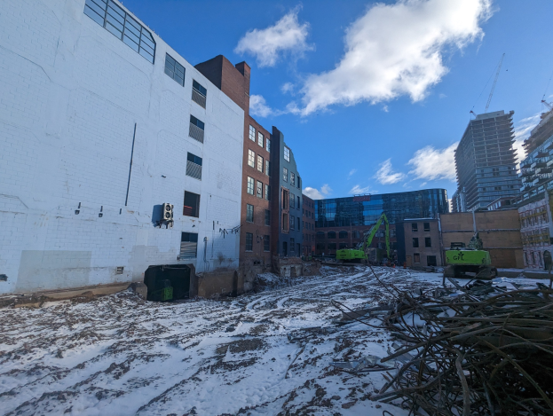 Großes Gebäude mit schneebedeckter Erde, umgeben von Fahrzeugen und Schutt, mit bewölktem Himmel und anderen Gebäuden im Hintergrund.