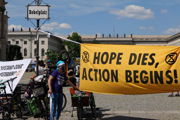 Eine Gruppe von Menschen steht vor einem Gebäude und hält ein gelbes Banner mit der Aufschrift "Hope Dies, Action Begins". In der Nähe stehen Fahrräder und ein Mast mit einer Tafel. Im Hintergrund sind Bäume und ein klarer blauer Himmel zu sehen.