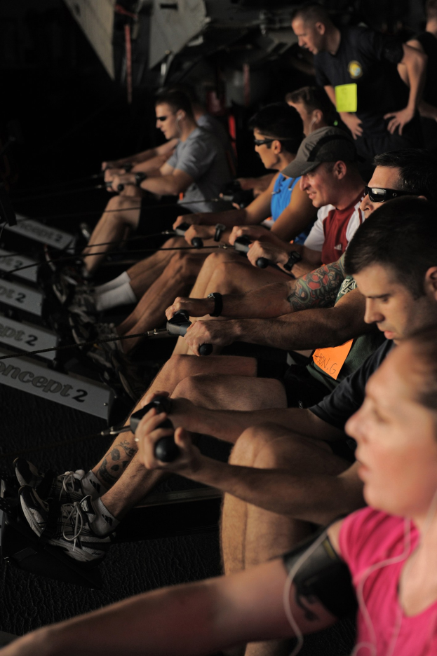 Eine Gruppe von Menschen, die an Rudermaschinen in einem Fitnessstudio trainieren, mit anderen im Hintergrund, die alle Sportschuhe tragen.