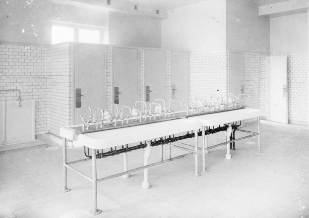 Schwarzes und weißes Foto einer öffentlichen Toilette mit einer Reihe von Waschbecken, Wasserhähnen, Wänden, Türen, Fenstern und einem gefliesten Boden, mit verschiedenen Gegenständen, die herumliegen.