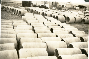 Ein Schwarz-Weiß-Foto eines großen Haufens von Betonrohren auf dem Boden mit ein paar Gebäuden im Hintergrund.