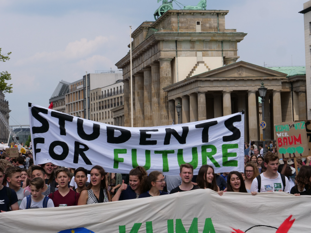 Gruppe von Schülern marschiert in Berlin mit einem buntfarbenen "Students for Future"-Schild vor einem Hintergrund aus Gebäuden, Bäumen und Himmel.