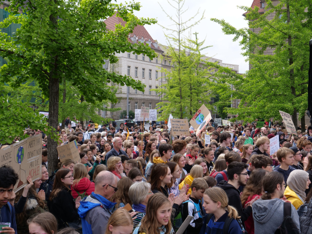 Eine große Menge von Menschen protestiert vor einem Gebäude in Berlin, mit Schildern, Bäumen, Fahrzeugen und dem Himmel.