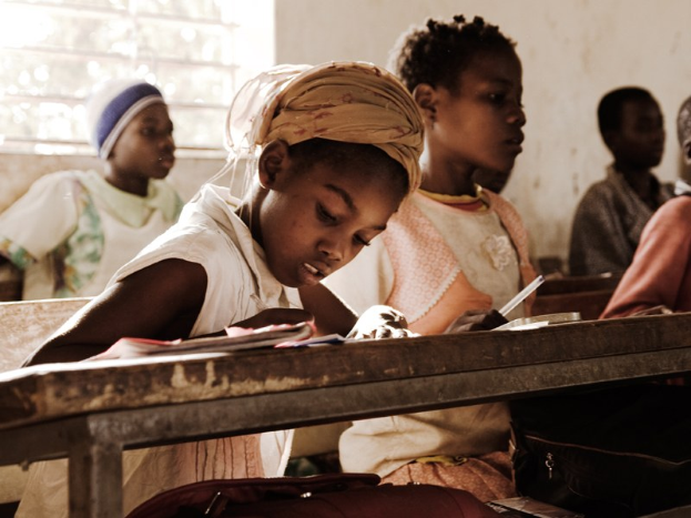 Eine Gruppe von Kindern sitzt an Tischen in einem Klassenzimmer, mit Büchern und Taschen auf den Bänken vor ihnen und einem Fenster im Hintergrund.