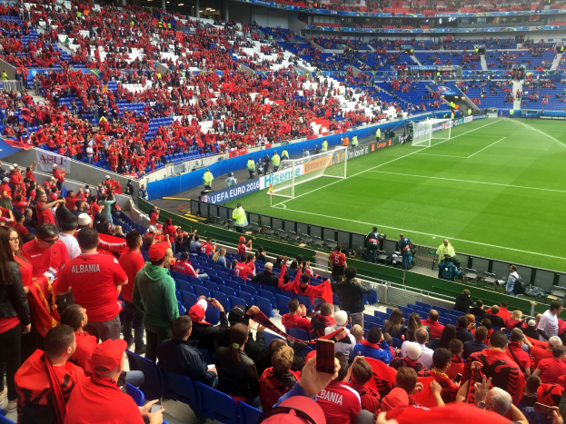 Eine große Menschenmenge in einem Stadion schaut einem Fussballspiel zu, mit Menschen, die auf Stühlen sitzen und stehen, Schildern, einem Torpfosten und Deckenleuchten.