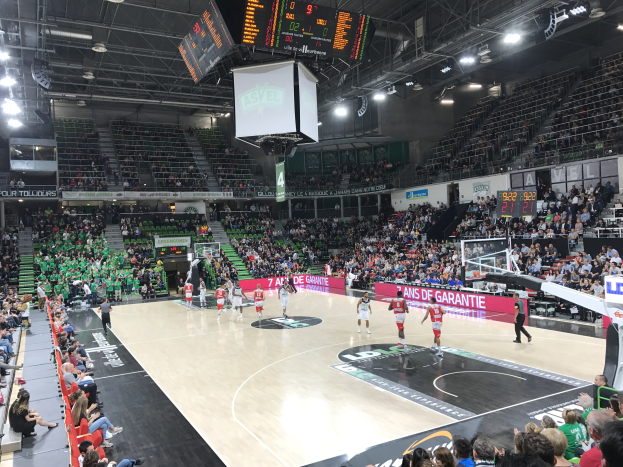 Ein Basketballspiel in einer großen Arena mit Zuschauern auf den Rängen, Anzeigetafeln mit "Asvel vs Galatasaray" und einer beleuchteten Decke mit einem Bildschirm über dem Spielfeld.